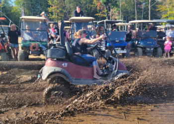 A golf cart driving through the mud with other golf carts watching from a distance.